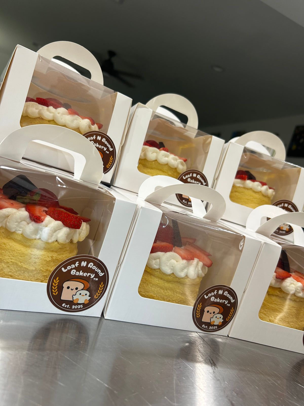 Several white bakery boxes with clear front windows and white handles, each containing a 4” cheesecake with cream, strawberries, arranged on a metallic counter. A round 'Loaf N Round Bakery' sticker is visible on the front of the boxes.