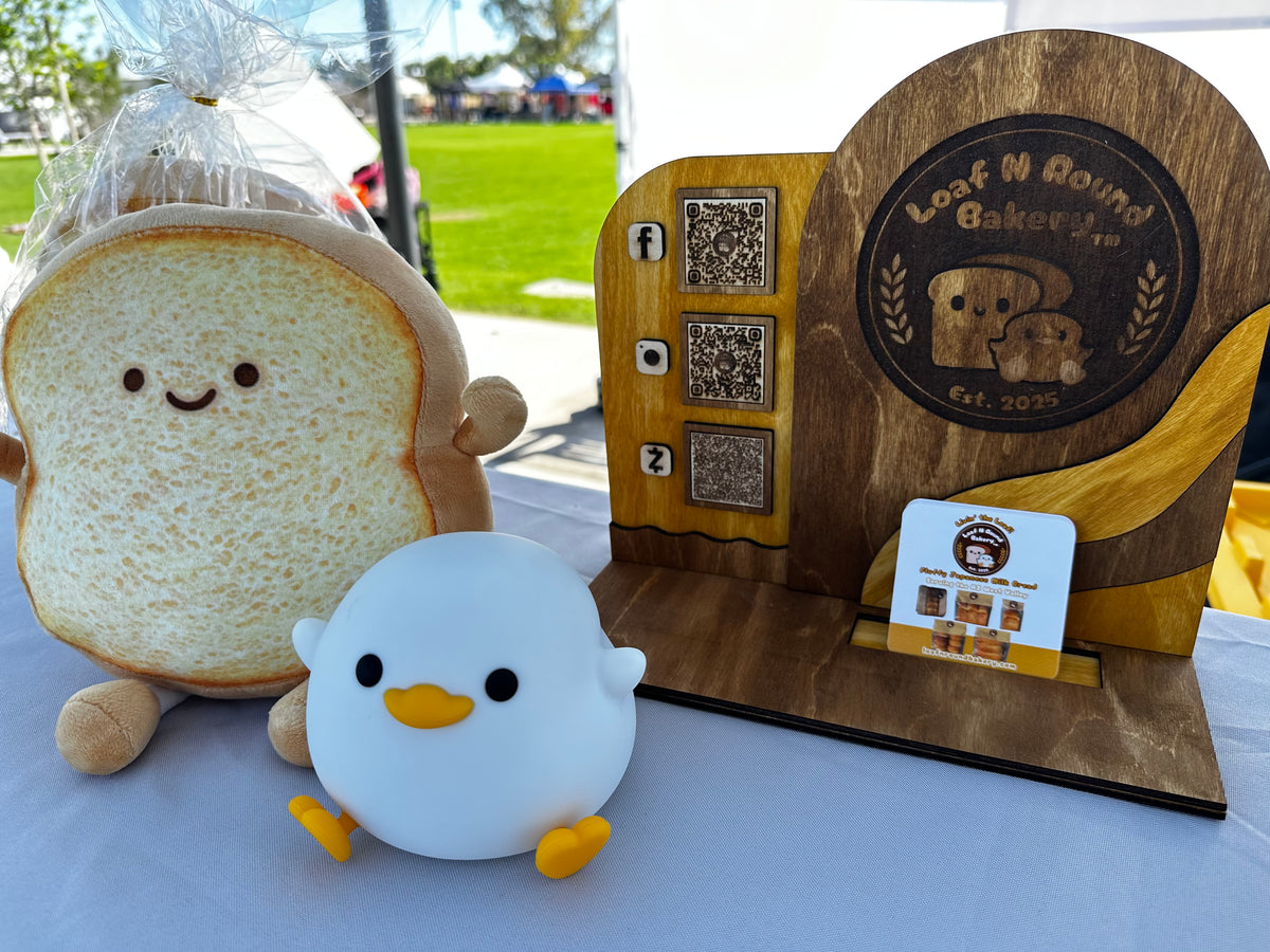 A wooden business plaque for 'Loaf N Round Bakery' displayed on a table next to a cute plush toy shaped like a slice of toast and a small white duck-shaped figurine.