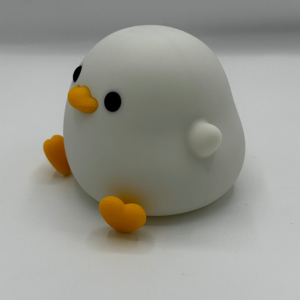 A cute, minimalist white duck figurine with a yellow beak, black eyes, and yellow feet, sitting on a plain light gray surface.